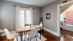 Un comedor con mesa de madera y sillas blancas, una ventana con cortinas blancas y un reloj en la pared.