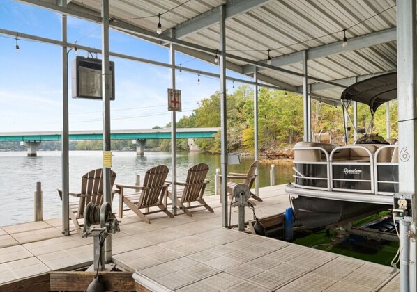 A covered dock with Adirondack chairs, a boat, and a view of a bridge over water.