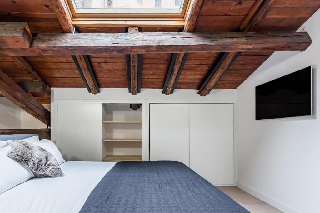 A bedroom with a skylight, wooden beams, a bed, a wardrobe, and a TV on the wall.