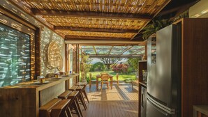 A kitchen with a wooden bar, a refrigerator, and a view of a garden. - Santi Sabai Ibira - Um Refúgio às Margens da Lagoa (Imbituba)