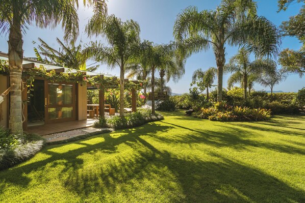 A well-maintained garden with palm trees, a wooden deck, and a clear sky. - Santi Sabai Ibira - Um Refúgio às Margens da Lagoa (Imbituba)