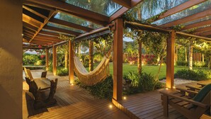 Una terraza de madera con una hamaca, sillas de madera y un techo de vidrio.