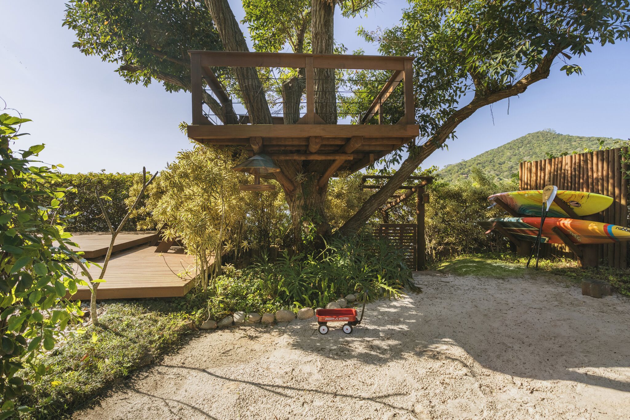 A wooden treehouse platform with a swing, surrounded by greenery and a sandy path.