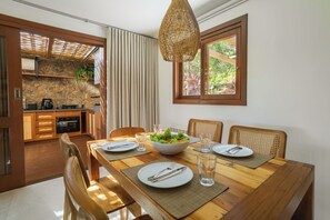 A dining area with a wooden table set for four, a bowl of salad, and a window with a view of greenery. - Santi Sabai Ibira - Um Refúgio às Margens da Lagoa (Imbituba)