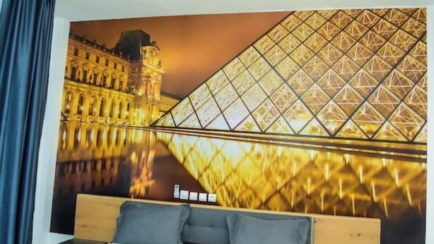 A hotel room with a large bed, a nightstand, and a wall mural of the Louvre's glass pyramid.