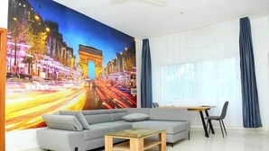 A modern living room with a large mural of the Arc de Triomphe, a grey sectional sofa, a glass-top coffee table, and a dining area with a small table and chairs.