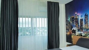 Une chambre d’hôtel avec un lit, une grande fenêtre ornée de rideaux légers et un tableau représentant un paysage urbain accroché au mur.