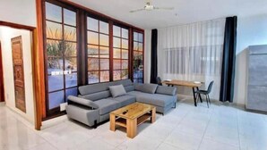 Un salon moderne avec un canapé d’angle gris, une table basse en bois et un coin repas comprenant une table et des chaises. De grandes fenêtres offrent une vue sur l’extérieur.