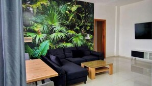 A modern living room with a black sofa, a wooden coffee table, and a large tropical-themed mural on the wall. - Jungle Sensation (Lomé)