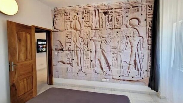 Una stanza con un ampio affresco raffigurante geroglifici e figure dell'antico Egitto, una porta in legno e una finestra con tende.