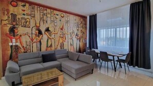 Un salon agrémenté d’une grande fresque murale sur le thème de l’Égypte, d’un canapé d’angle gris, d’une petite table basse en bois et d’un coin repas avec une table et des chaises.