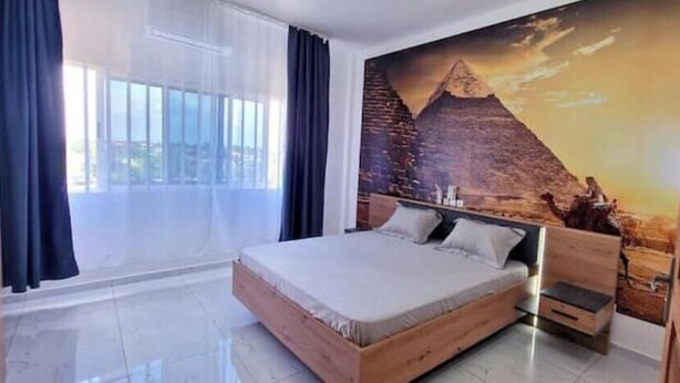 Une chambre avec un grand lit, une petite table de chevet, une fresque murale représentant une pyramide et un balcon avec des rideaux.