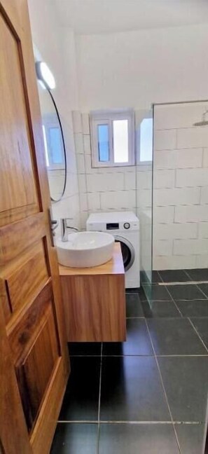 A compact bathroom with a wooden vanity, a sink, a washing machine, and a tiled floor. - Voltiges.. (Lomé)