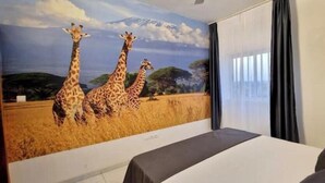 Ett hotellrum med en stor väggmålning som föreställer giraffer, en säng och ett fönster med gardiner.
