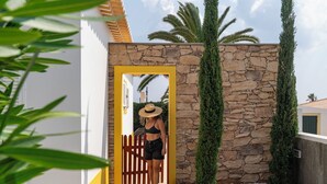 Una persona entra a un edificio por una puerta amarilla, con un muro de piedra y altos árboles verdes al fondo.