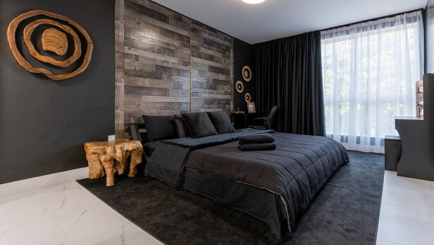 Ein modernes Schlafzimmer mit einem großen Bett, dunkler Bettwäsche, einer Wand mit Holzpaneelen und Blick auf Bäume durch transparente Vorhänge.