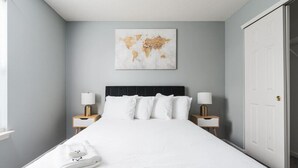 A neatly made bed with white linens, two bedside tables with lamps, a world map artwork on the wall, and a closed door. - Cozy Home Plainfield Indy (Plainfield)