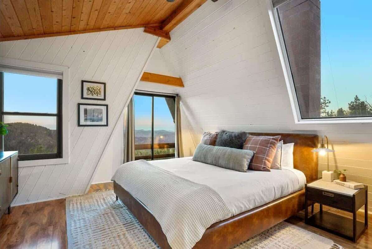 Een slaapkamer met een schuine plafond, een groot bed met veel kussens, een nachtkastje met lamp en een raam met uitzicht op de omgeving.