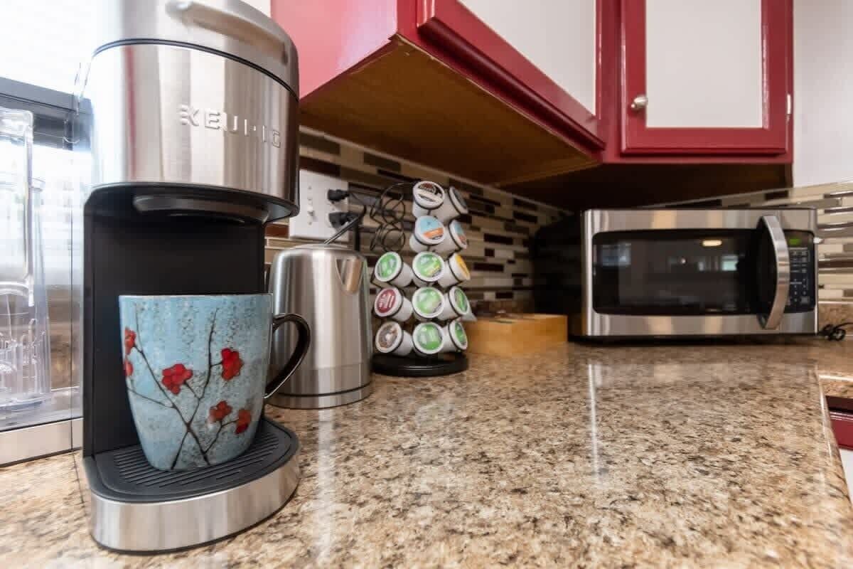 Eine Küche mit einer Keurig-Kaffeemaschine, einer Tasse mit Blumenmuster, einem Stapel bunter Tassen und einer Mikrowelle.