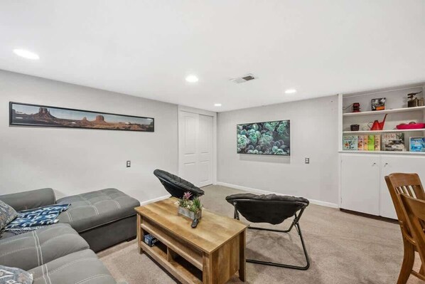 A living room with a gray sectional sofa, a wooden coffee table, and a black folding chair. There are two framed pictures on the walls, a white door, and a white shelving unit with various items.
