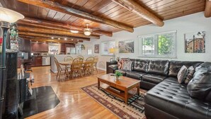 A spacious living room with a wooden ceiling, a black leather sofa, a wooden coffee table, and a dining area with wooden chairs and a table. - Pickleball Retreat | Hot Tub & Games (Denver)