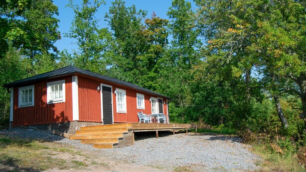 Eine rote Holzhütte mit einer Holzdielen-Terrasse und einem Kiesweg, der davor führt, umgeben von Bäumen.