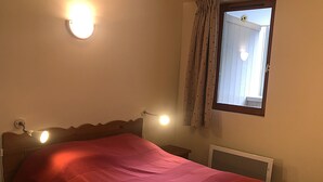 A single bed with a red bedspread, two bedside lamps, a window with curtains, and a wall-mounted light fixture. - Terrasses F 102 - PARC NAT. VANOISE appart. 4 pers (Val Cenis)