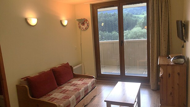 A room with a sofa, a table, and a wooden cabinet. There is a sliding glass door leading to an outdoor area with a view of trees. - Terrasses F 102 - PARC NAT. VANOISE appart. 4 pers (Val Cenis)
