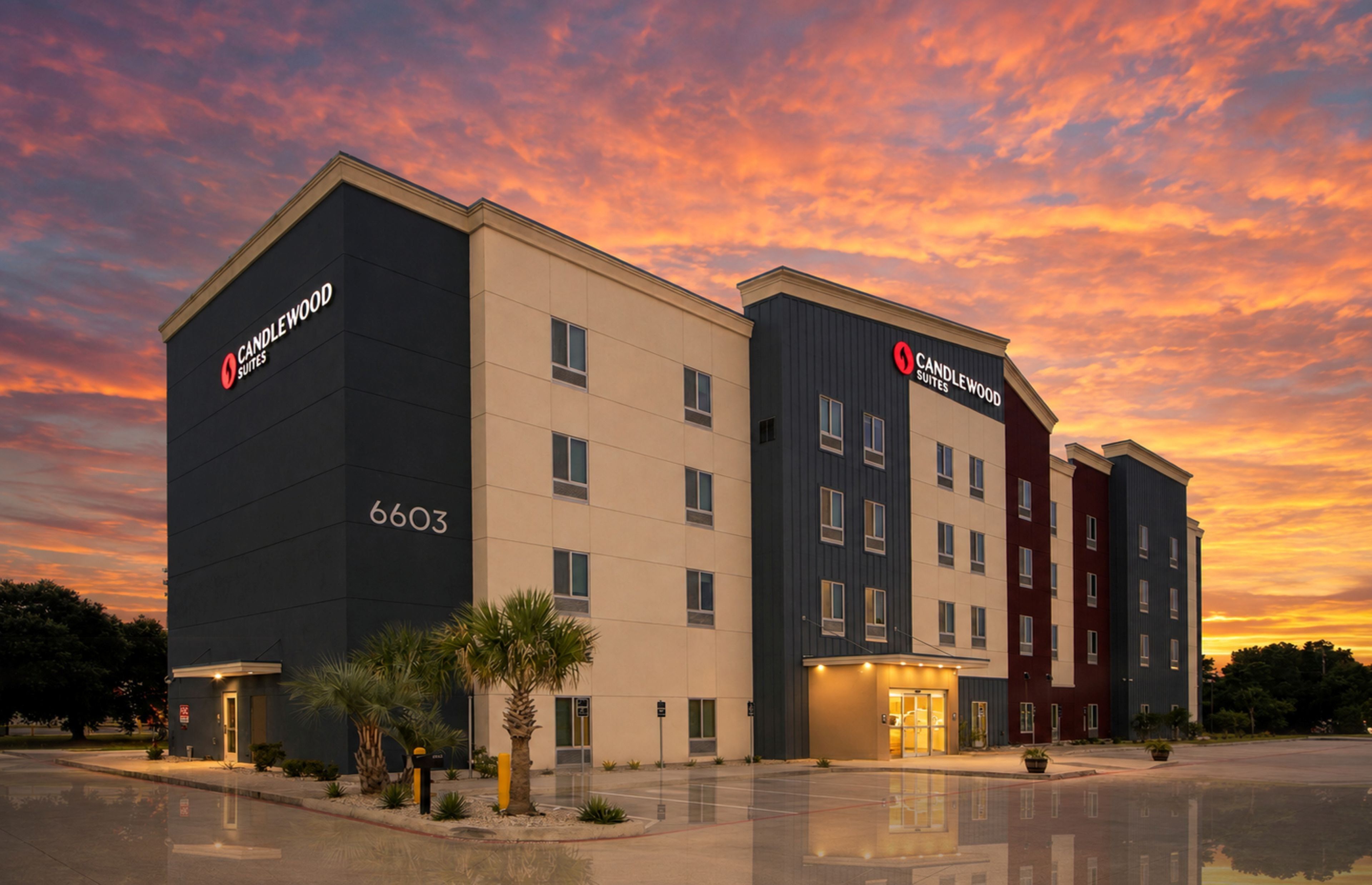 Un hotel con diseño moderno, que destaca un letrero grande de 'CANDLEWOOD SUITES', un techo plano y un estacionamiento reflectante.