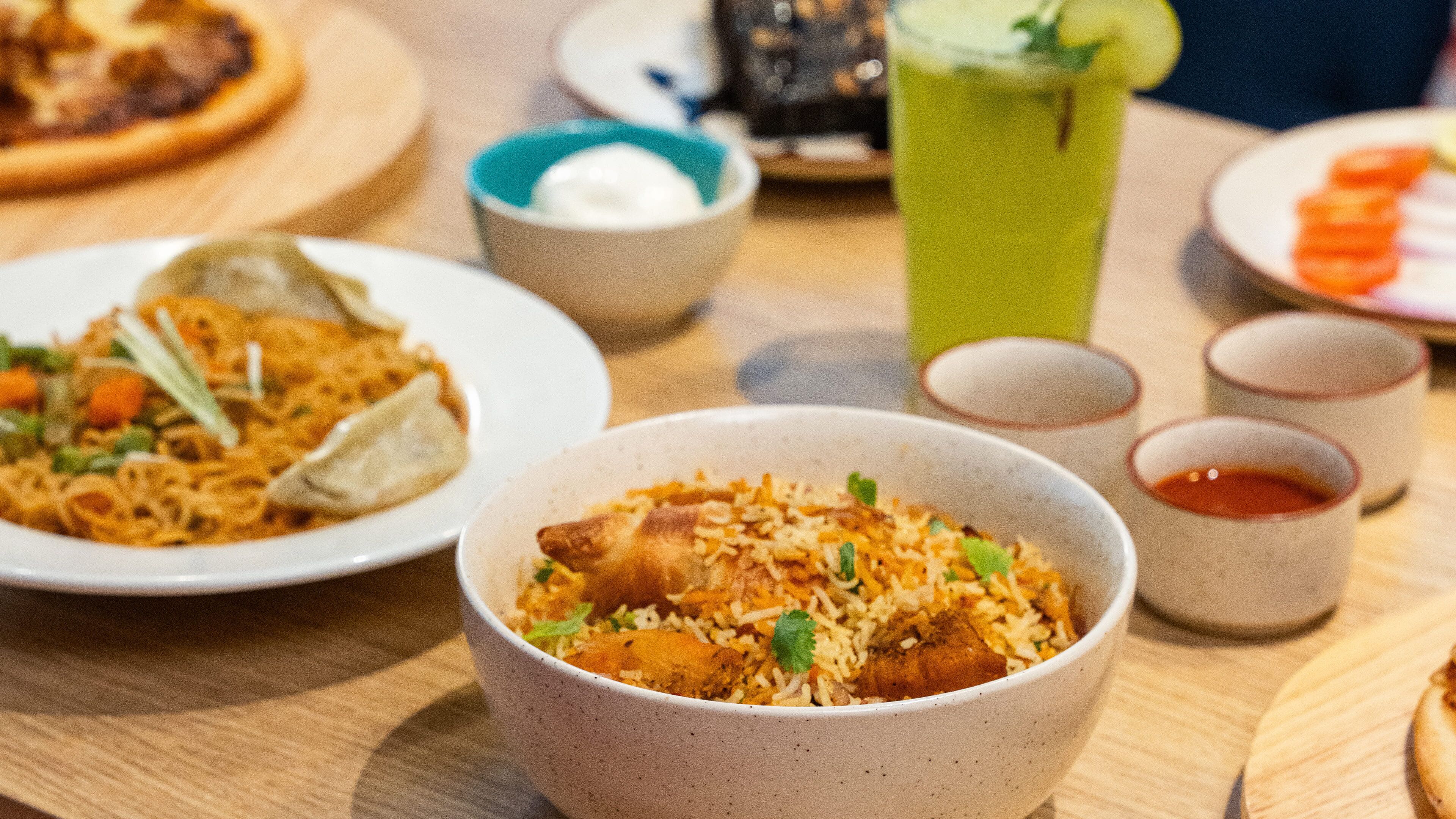 A dining table with various dishes including a bowl of biryani, a plate of noodles, and a green drink.