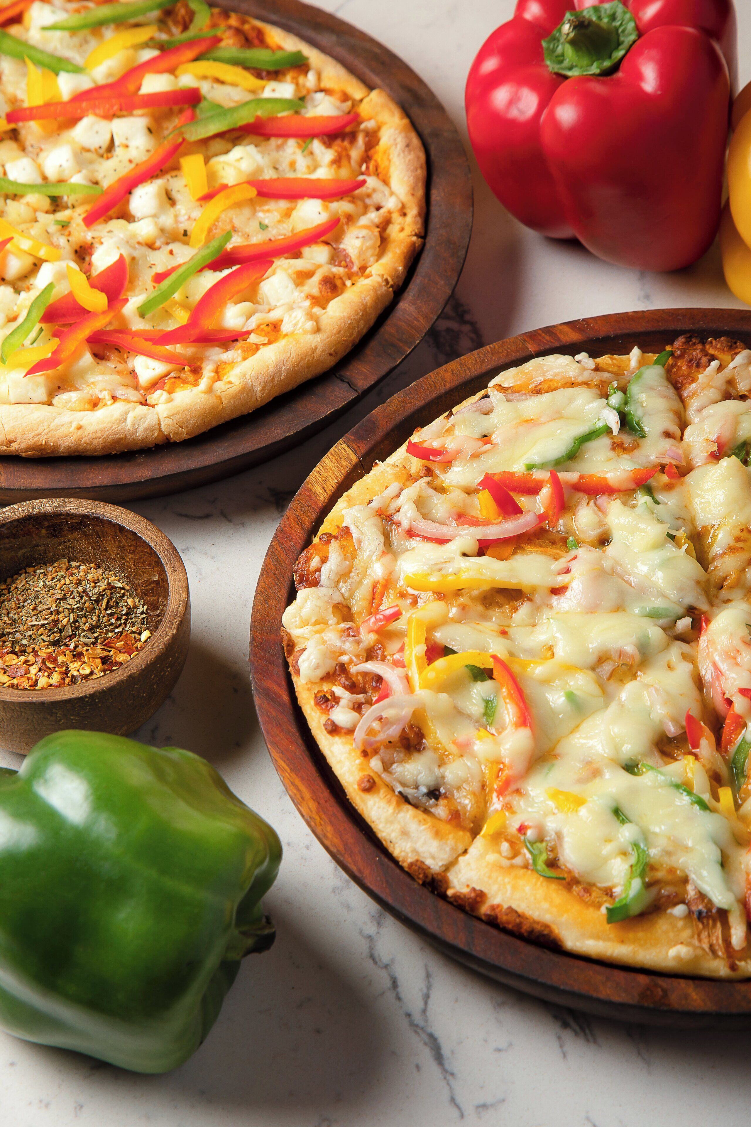 Two pizzas on wooden boards with various toppings, surrounded by whole bell peppers.