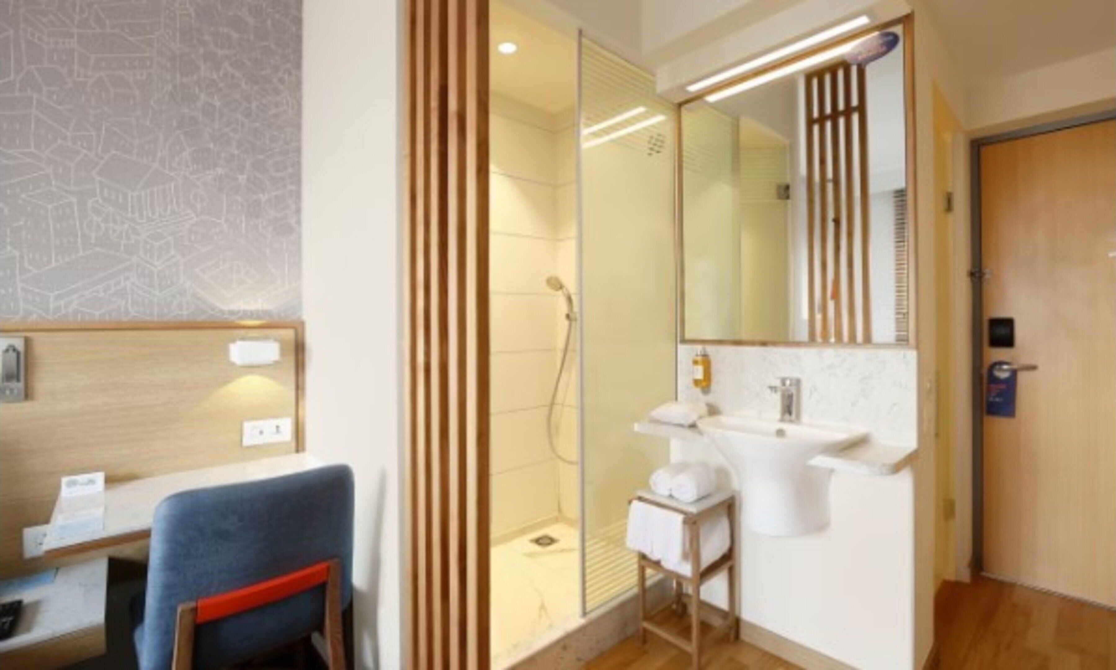 A modern bathroom with a shower, a sink, and a mirror. There is a wooden door and a small table with a towel.