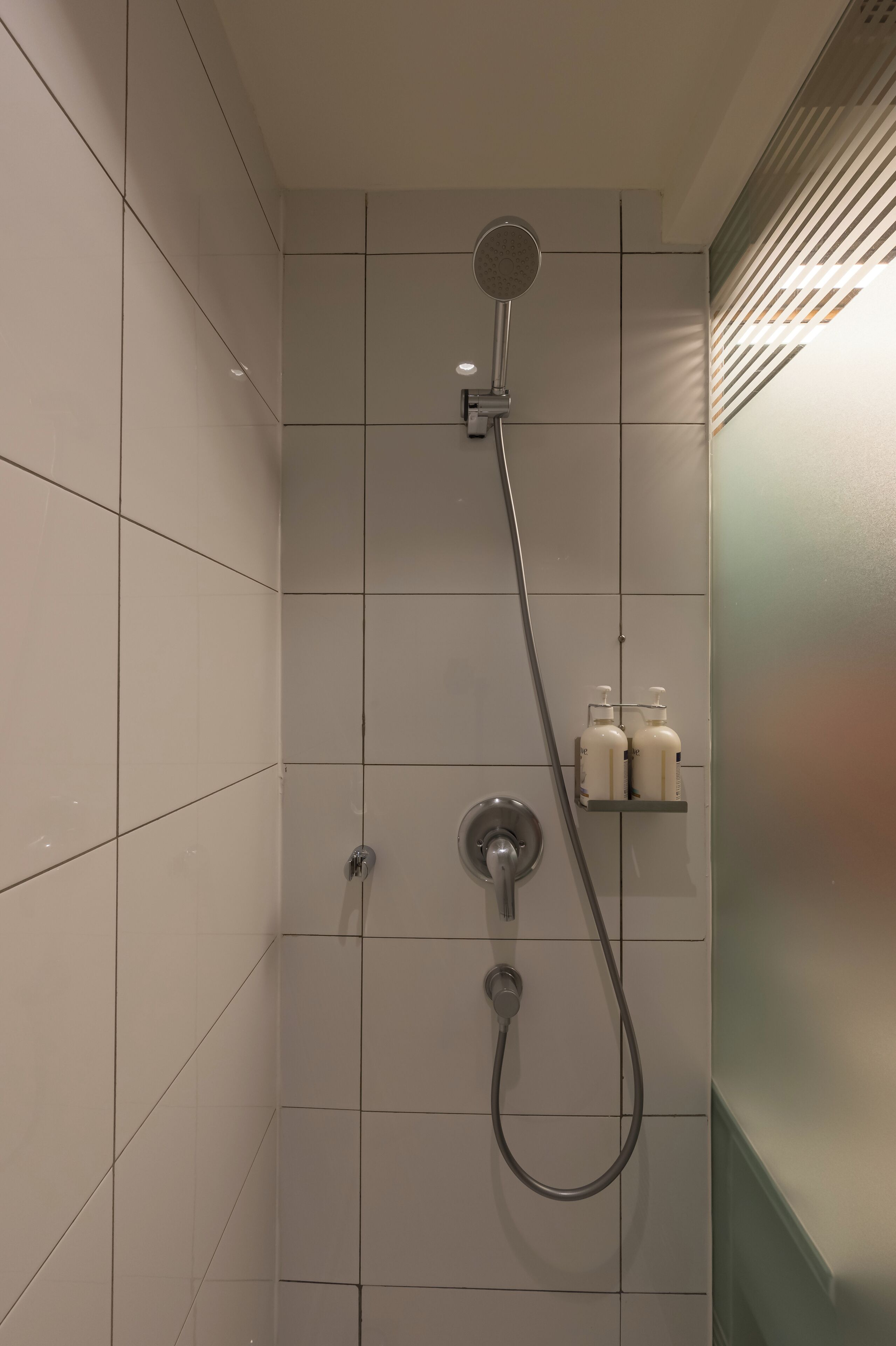 A shower with a handheld showerhead and a wall-mounted faucet. There are two bottles on a shelf above the shower.