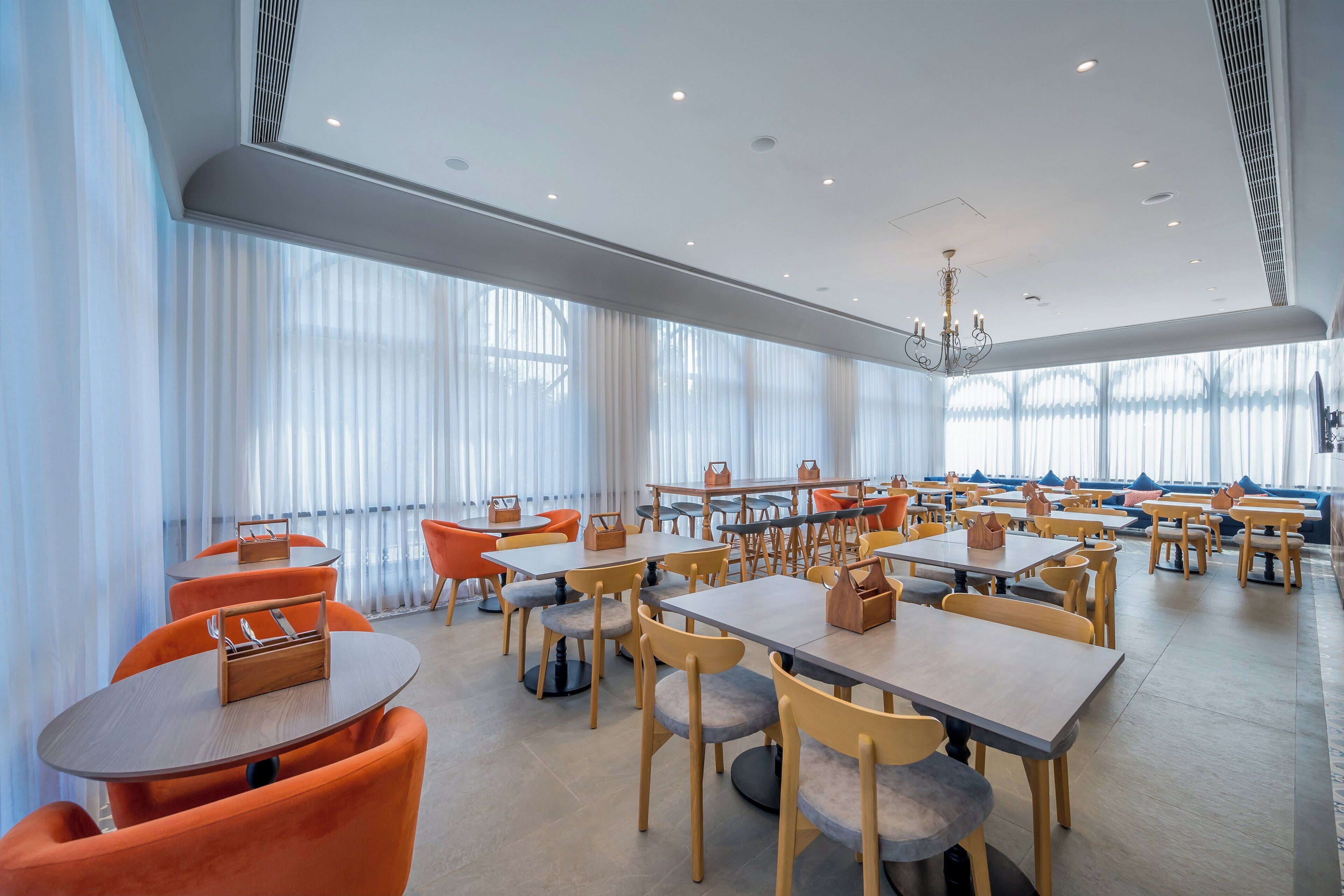 Un comedor amplio con varias mesas y sillas, ventanas grandes con cortinas translúcidas y una lámpara moderna.