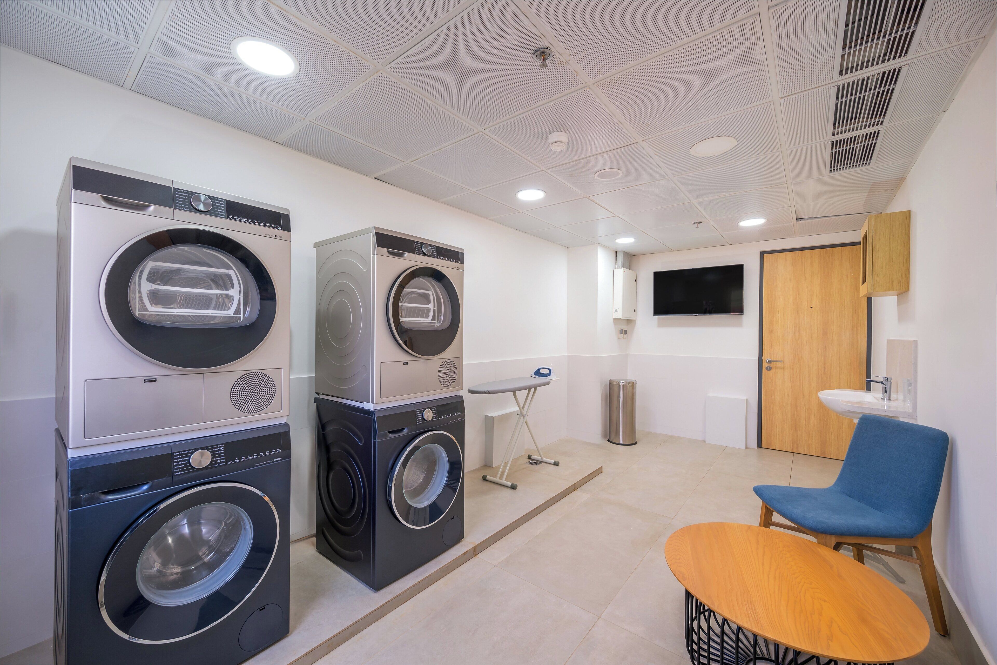 A laundry room with stacked washing machines and dryers, a small table, and a blue chair.