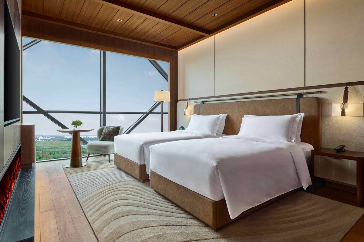 A modern hotel room with two beds, a large window offering a city view, a fireplace, and a small table with a lamp.