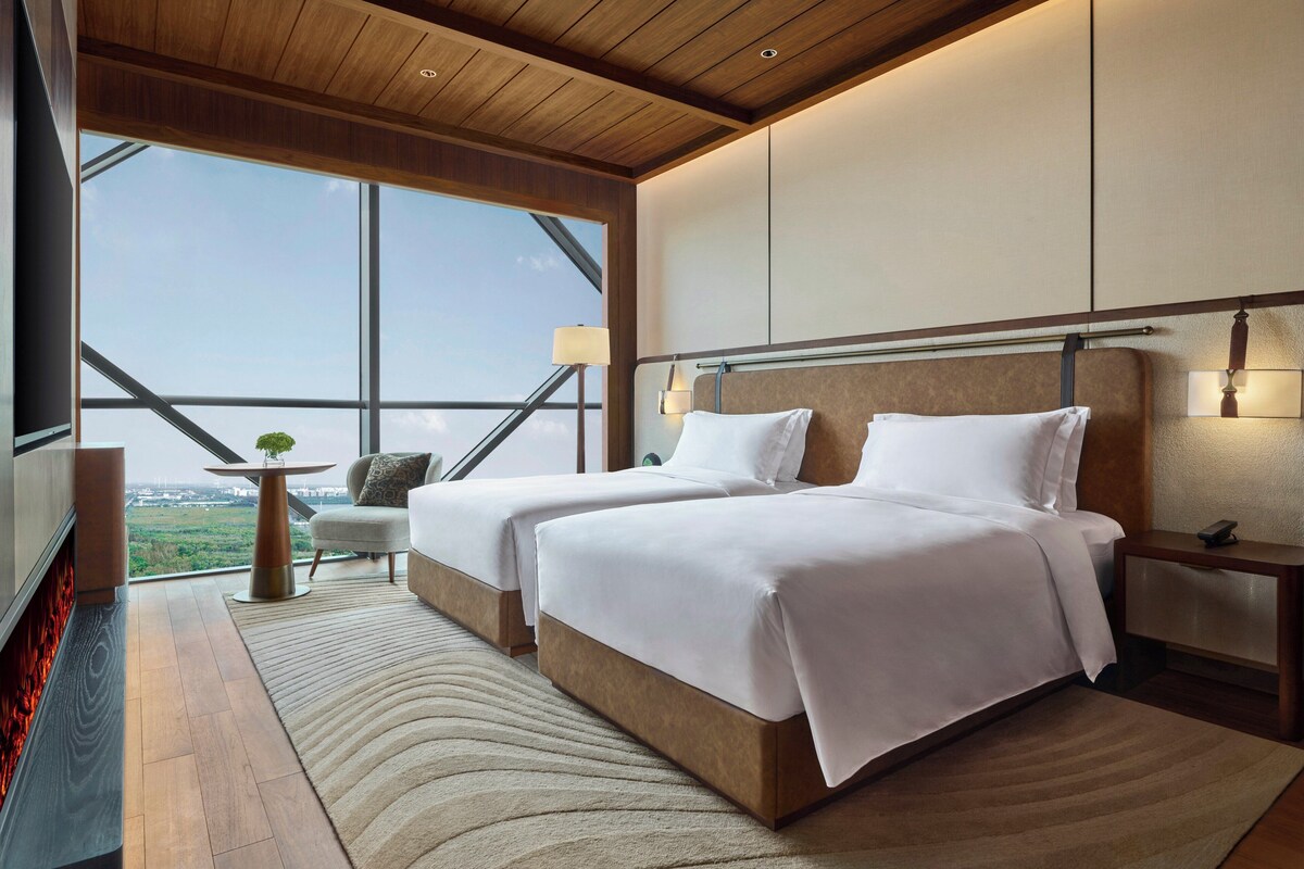 A modern hotel room with two beds, a fireplace, a large window with a view, and a small table with a lamp.