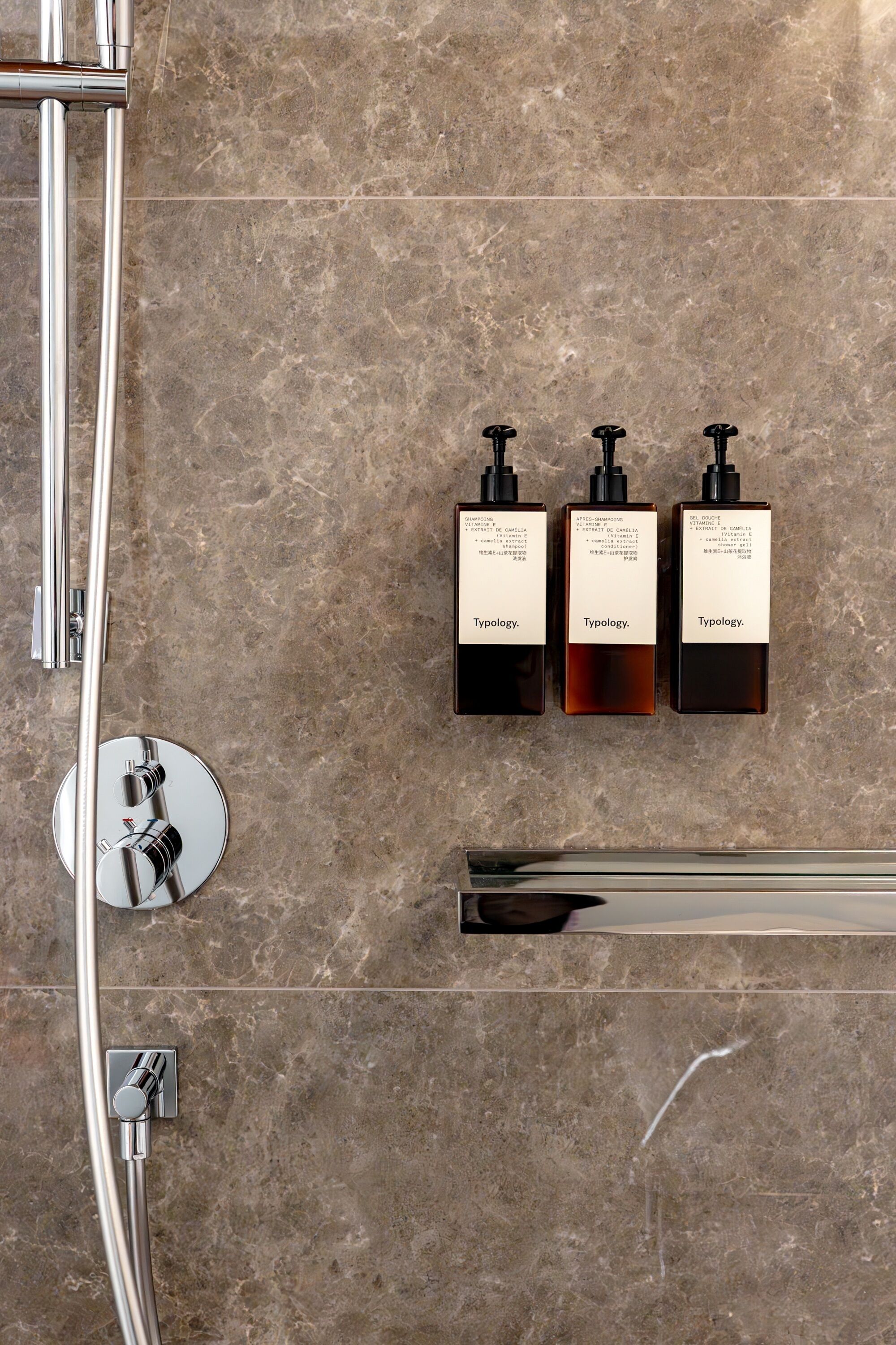 A bathroom with a marble wall, a chrome shower head, and three bottles of Typology body wash.