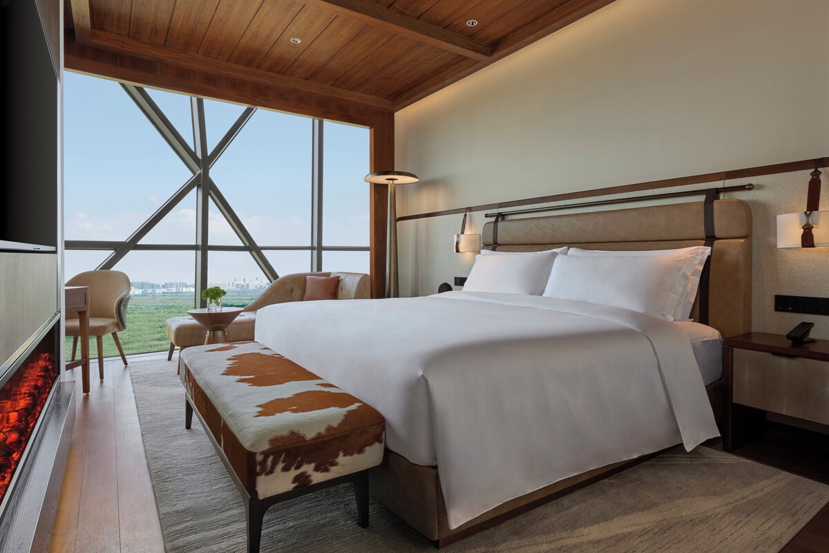 A modern hotel room with a large bed, a wooden ceiling, a view of the city, and a small seating area.