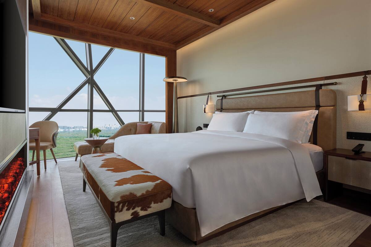 A modern hotel room with a large bed, a wooden ceiling, a view of the city, and a small seating area.