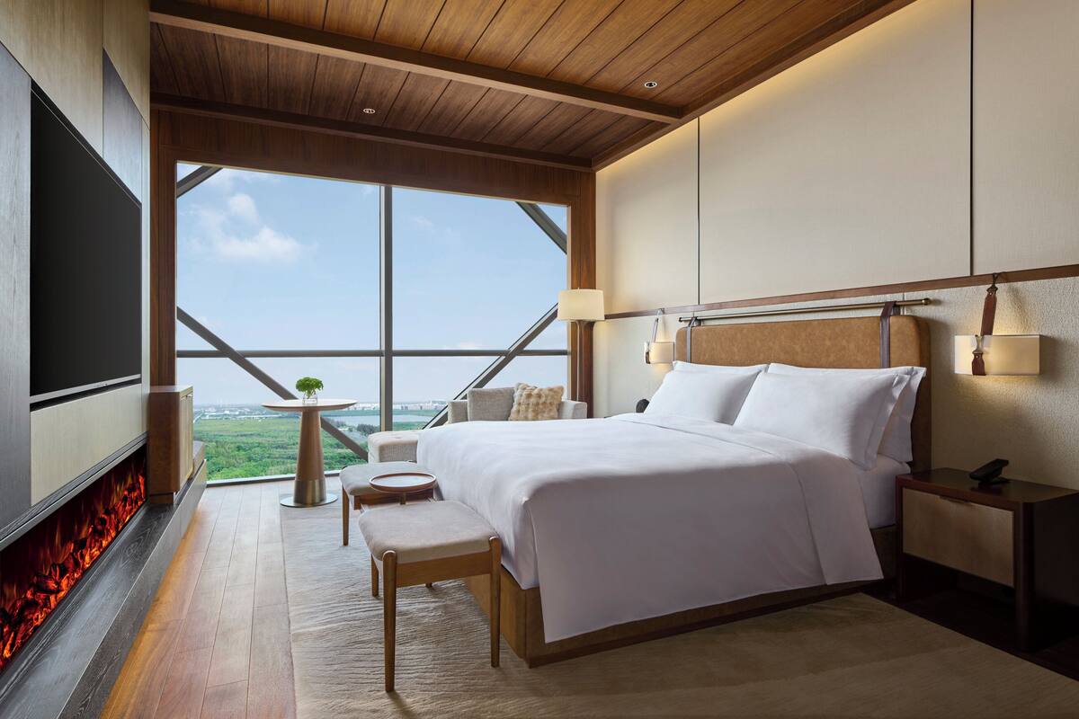 A modern hotel room with a large bed, a TV, a fireplace, and a view of the landscape.