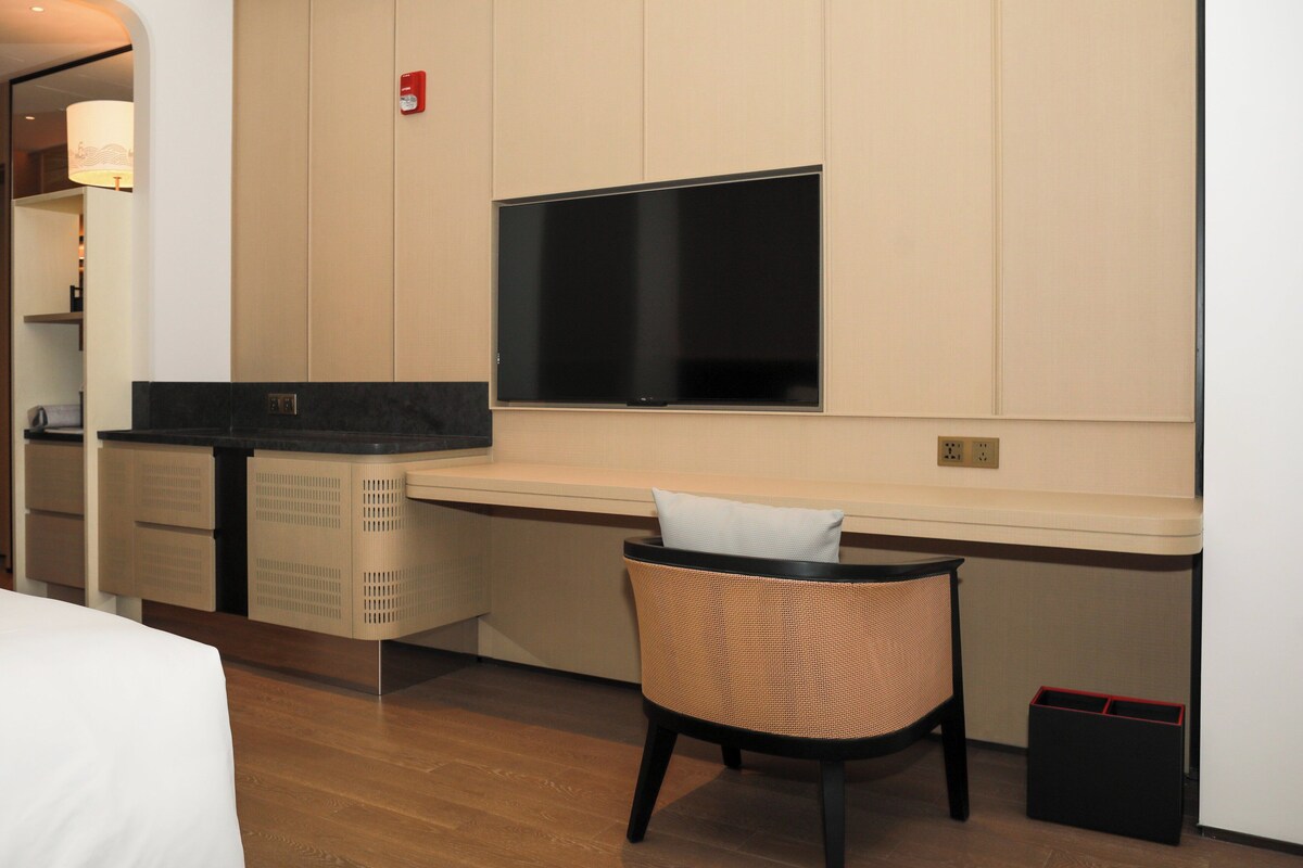 A hotel room with a bed, a desk with a flat-screen TV, a chair, and a laundry basket.