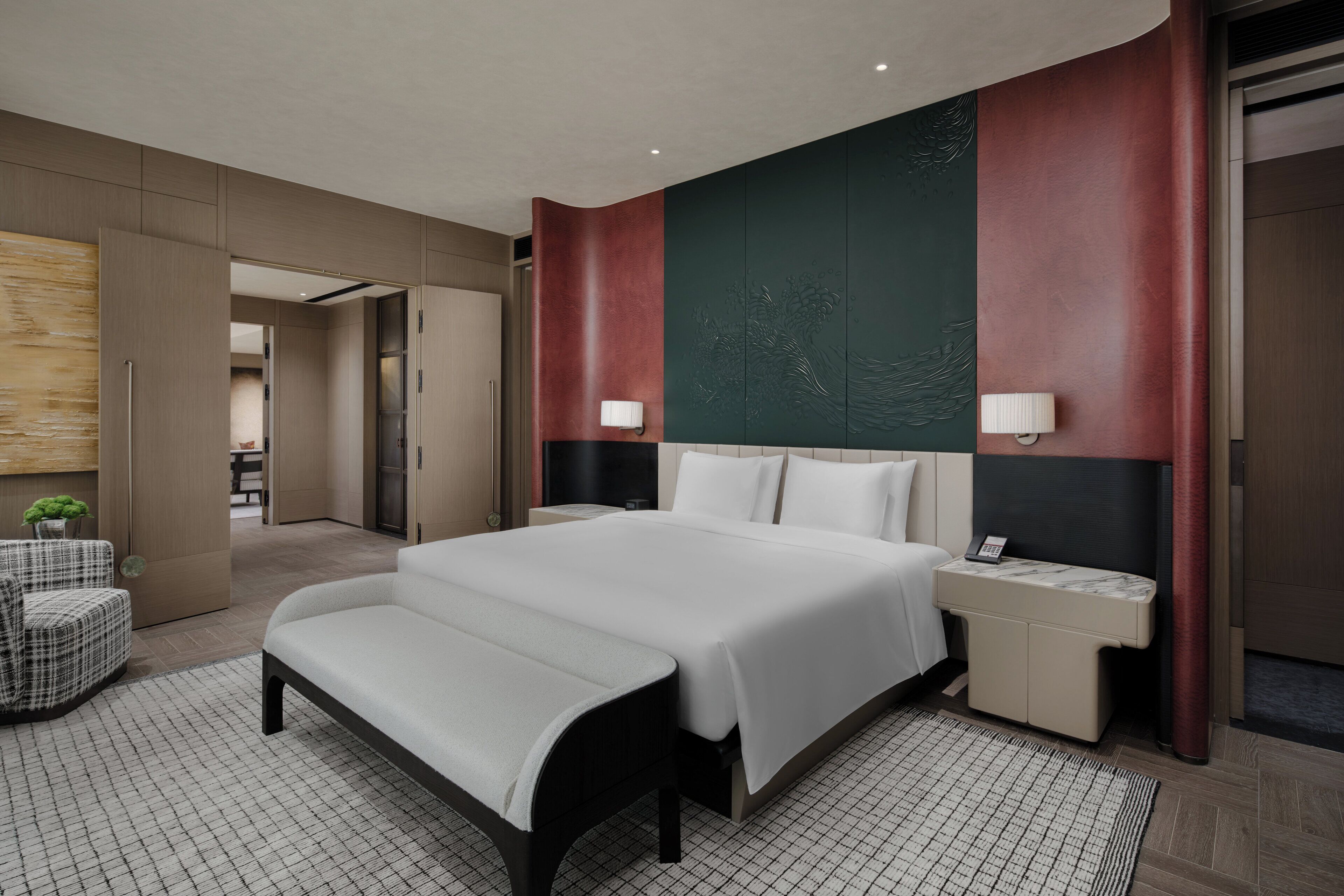 A modern hotel room with a large bed, bedside tables, a bench, and a view of the hallway.