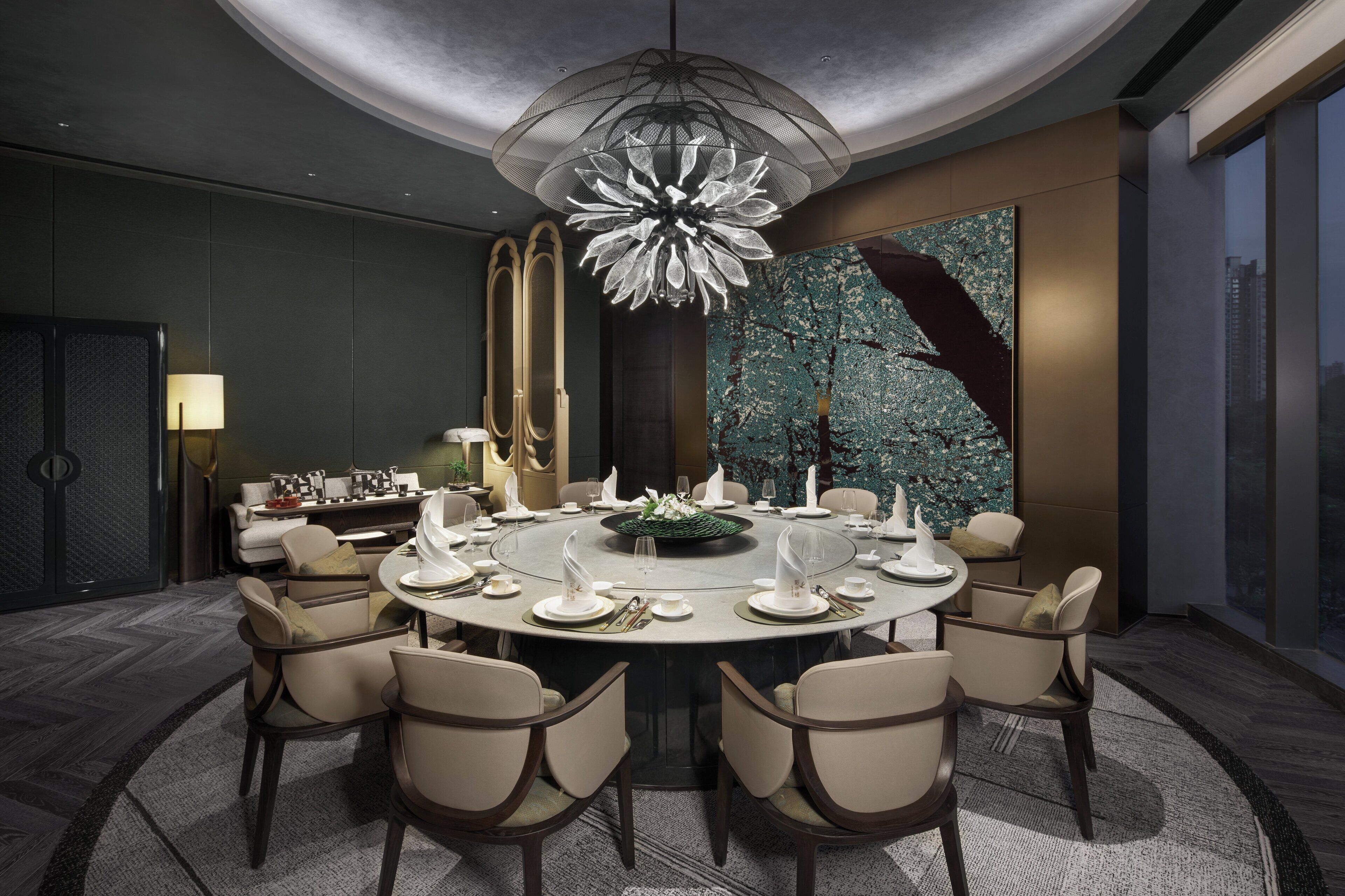 Une salle à manger moderne avec une table ronde dressée pour six personnes, entourée de chaises, d’un grand lustre et offrant une vue sur le paysage urbain.