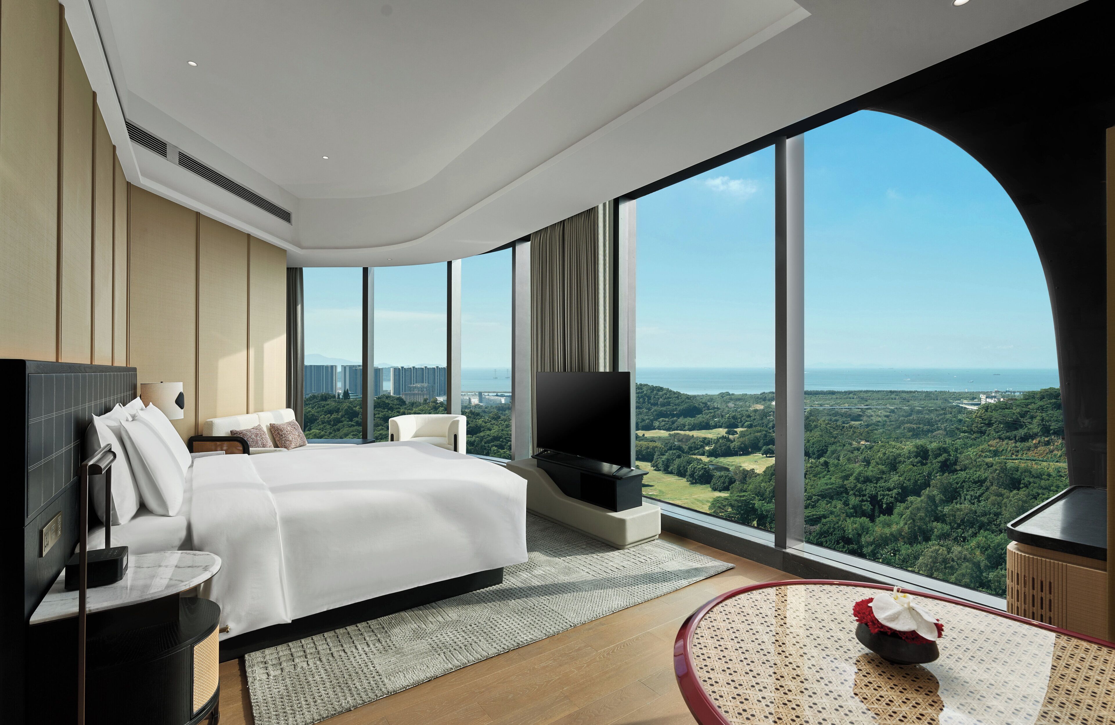 Una habitación de hotel moderna con una cama grande, un televisor de pantalla plana, una mesa de comedor y vistas panorámicas de la ciudad y zonas verdes.
