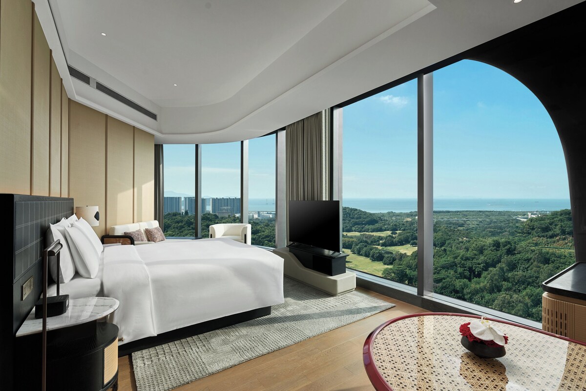A modern hotel room with a large bed, a flat-screen TV, a dining table, and a panoramic view of the city and greenery.