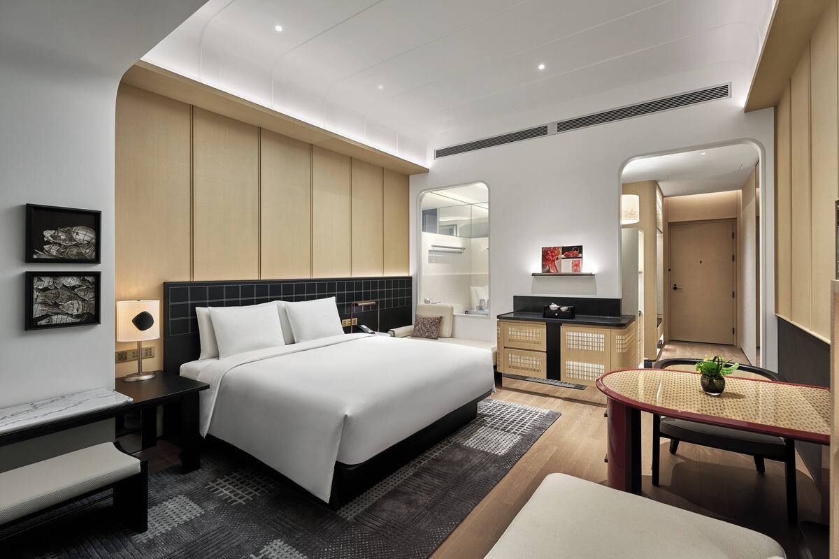 A modern hotel room with a large bed, bedside tables, a small desk, a chair, a round table with a plant, and a mirror.