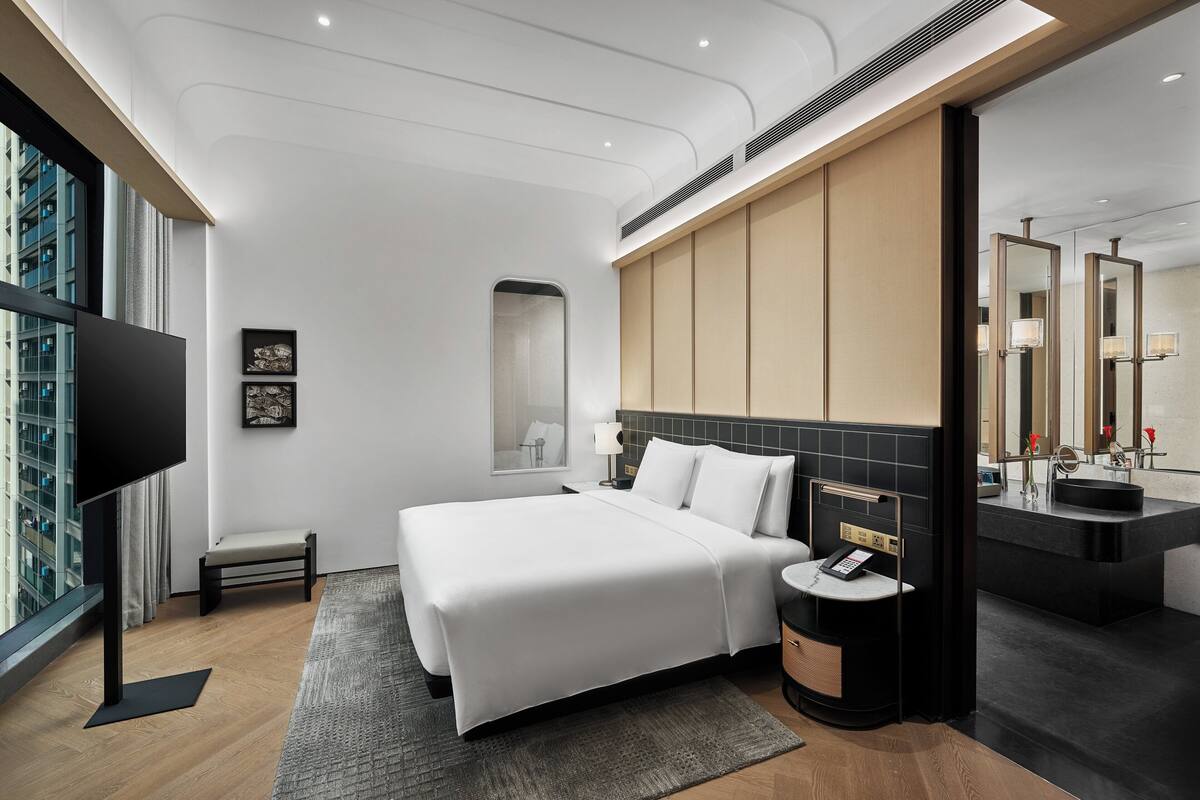A modern hotel room with a large bed, a bedside table with a phone, a small seating area, a large window, and a bathroom with a mirror and sink.