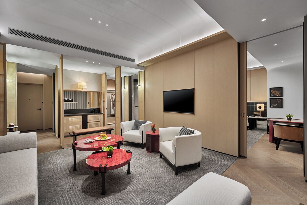 A modern hotel room with a sofa, two armchairs, a coffee table, a TV, and a kitchen area.