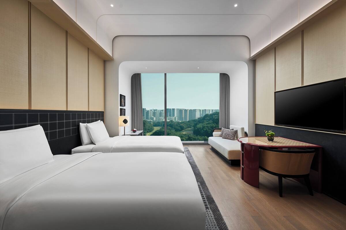A modern hotel room with two beds, a sofa, a desk, and a large TV.